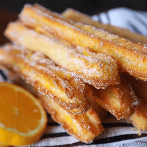 A beautiful stack of citrus zest churros. They are golden yellow, covered in sugar and garnished with orange peel with orange slices on the side